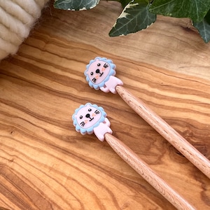 May include: Two wooden knitting needles with pink and blue lion-shaped tops. The lions have black eyes and a smiling mouth.