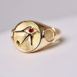 May include: A gold ring with an oval-shaped design featuring the Eye of Horus with a red gemstone in the center.