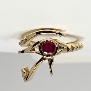 May include: Gold ring with a red ruby stone set in the center, shaped like the Eye of Horus, an ancient Egyptian symbol of protection.