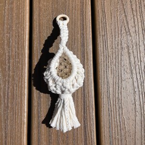 Pode incluir: Um pequeno suporte de plantas em macramé na cor branco sujo com uma estrutura de cesto atado, um anel de madeira circular no topo para pendurar e uma borla de franjas decorativa na parte inferior com uma abertura central visível.