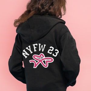 May include: Black zip-up hoodie with a white "NYFW 23" graphic and a pink star design on the back.