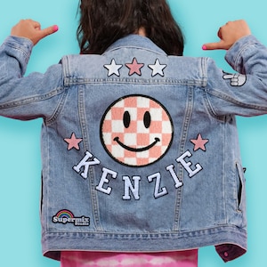 May include: A blue denim jacket with patches. The patches include a pink and white checkered smiley face, three stars, the word "KENZIE" in white letters, and a rainbow logo that says "Supermix Studio".