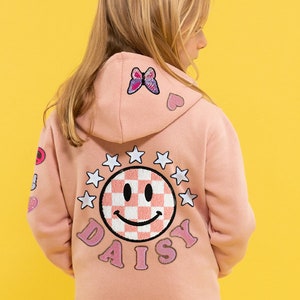 May include: A pink zip-up hoodie with a checkered smiley face and the word "DAISY" embroidered on the back. The hoodie has several colorful patches, including a butterfly, a heart, and a star.