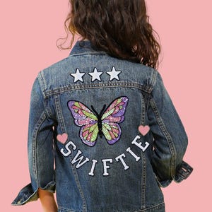 May include: A blue denim jacket with a butterfly and star embellishments on the back. The jacket has the word "SWIFTIE" written in white letters on the back.