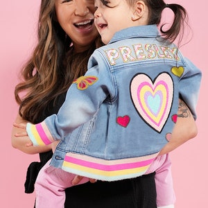 May include: A woman holding a young girl wearing a light blue denim jacket with colorful patches. The jacket has a rainbow heart patch on the back and the name "PRESLEY" written in colorful letters. The girl is wearing pink pants and white sneakers.