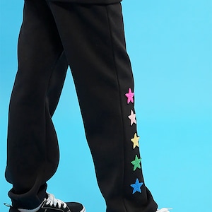May include: Black sweatpants with colorful star appliques on the side. The sweatpants are paired with black sneakers.