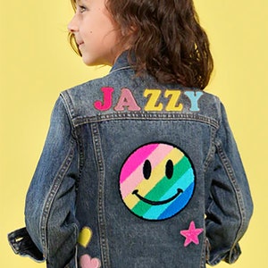 May include: A blue denim jacket with colorful patches, including a rainbow smiley face, a star, and hearts. The word "JAZZY" is spelled out in colorful letters on the back.