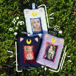 May include: Three colorful photo frames with a crescent moon and cat ears. The frames are made of yarn and have a soft, fuzzy texture. Each frame has a different color: white, purple, and black. The frames are decorated with a yellow crescent moon and pink cat ears. The frames are all empty and ready to be filled with photos.