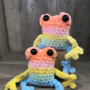 May include: Two crocheted rainbow frog plushies. The larger frog is sitting on a brown box and has yellow arms and legs. The smaller frog is sitting in front of the larger frog and has blue legs and yellow arms.