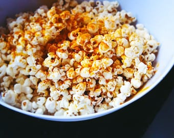 Może przedstawiać: Biała miska wypełniona popcornem, doprawionym żywą pomarańczową przyprawą. Ziarna popcornu są puszne i równomiernie pokryte, tworząc atrakcyjną wizualnie przekąskę. Miska stoi na ciemnym tle.