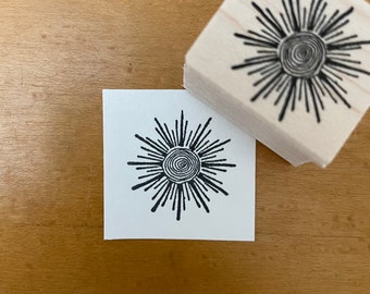 May include: A black and white rubber stamp with a design of a sun with a spiral in the center. The sun has many rays coming out from the center.