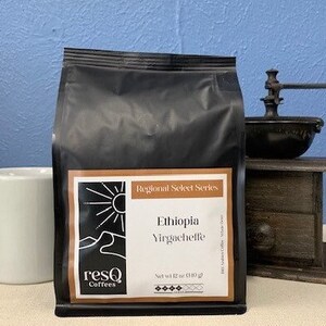 Puede incluir: Una bolsa negra de granos de café con una etiqueta marrón que dice "Regional Select Series Ethiopia Yirgacheffe" y "ResQ Coffees". La bolsa está sobre una mesa junto a un molinillo de café de madera.