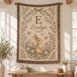 May include: A woven wall tapestry featuring a cream background with a central oval floral wreath tied with a green bow. Inside the wreath is the letter E, the names Evelyn Grace, and a brown rabbit surrounded by daisies and butterflies.