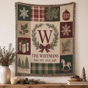 May include: A woven throw blanket featuring a central wreath with the letter W, the text THE WHITMANS and EST. 2025. The design includes panels with snowflakes, plaid patterns, pine branches, a gift box, and a rocking horse in red, green, and tan.