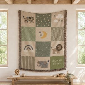May include: A woven throw blanket featuring a grid pattern with illustrations of a tiger, a crescent moon, stars, clouds, a rainbow, a lion, and an elephant. The blanket includes the text OLIVER SMITH and has a fringed edge in muted green and tan.
