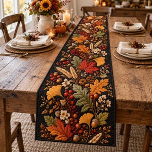 May include: A black quilted table runner featuring an embroidered autumn pattern of colorful maple leaves, ginkgo leaves, wheat stalks, berries, and acorns. The runner is centered on a wooden dining table with white napkins and place settings.