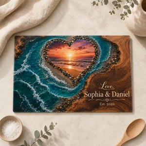 May include: A rectangular glass cutting board featuring a heart-shaped cutout design filled with a sunset ocean scene. The board displays the text Love, Sophia & Daniel, and Est. 2026. The surrounding area shows a sandy beach and turquoise waves.