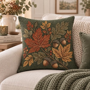 May include: A square throw pillow with a dark green textured fabric base featuring a woven pattern of autumn leaves in shades of orange, red, and gold, along with brown acorns and small red berries arranged in a central botanical cluster.