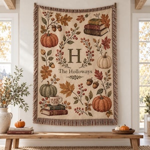 May include: A woven throw blanket featuring a cream background with a fall-themed pattern of pumpkins, acorns, autumn leaves, and stacked books. The center displays a large letter H and the text The Holloways. The blanket has fringed edges.