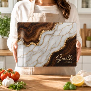 May include: A rectangular cutting board featuring a split design of dark brown wood grain and white marble patterns, separated by a jagged gold metallic vein. The text Smith and EST. 2026 is printed in gold script and block lettering on the lower right.
