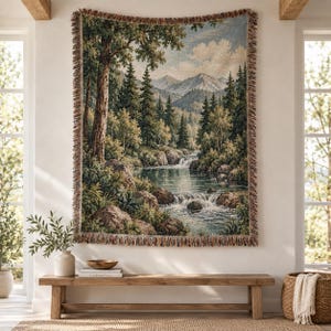 May include: A woven wall tapestry featuring a detailed landscape scene of a flowing river with small waterfalls, surrounded by dense evergreen trees, rocky terrain, and distant mountains under a cloudy sky, finished with a fringed edge border.