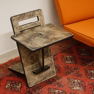 May include: A dark wood folding stool with a black metal bar across the top. The stool is sitting on a red and cream patterned rug.