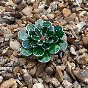 May include: A handcrafted, stained glass flower with green petals and a brown center. The flower is surrounded by a silver metal frame. The image is taken outdoors, with the flower resting on a bed of small rocks.