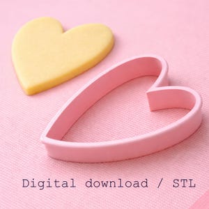 Puede incluir: Un cortador de galletas rosa en forma de coraz&oacute;n junto a una galleta en forma de coraz&oacute;n sobre una superficie rosa. La galleta es amarilla. El texto "Digital download / STL" est&aacute; en la parte inferior de la imagen.