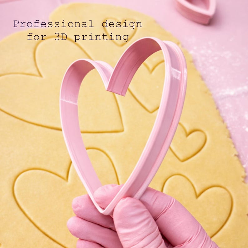Puede incluir: Un cortador de galletas rosa en forma de coraz&oacute;n sostenido sobre masa con huellas de coraz&oacute;n. El texto "Professional design for 3D printing" es visible. El cortador es de pl&aacute;stico y se utiliza para hornear.
