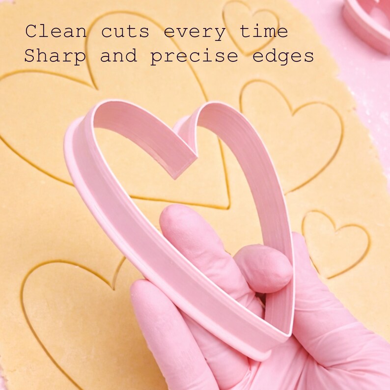 Puede incluir: Un cortador de galletas rosa en forma de coraz&oacute;n sobre masa con contornos de coraz&oacute;n. La imagen incluye el texto "Clean cuts every time" y "Sharp and precise edges". El cortador es sostenido por una persona con guantes rosas.