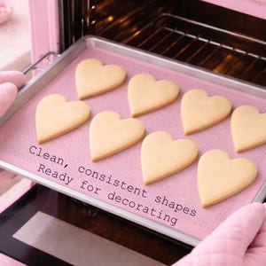 Puede incluir: Una bandeja para hornear con galletas en forma de coraz&oacute;n se saca de un horno rosa. Las galletas son de color amarillo claro y est&aacute;n sobre una bandeja rosa. El texto en la bandeja dice: "Formas limpias y consistentes, listas para decorar".