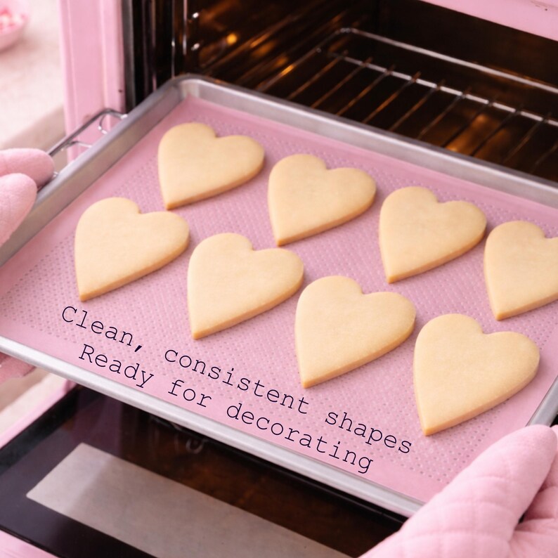 Puede incluir: Una bandeja para hornear con galletas en forma de coraz&oacute;n se saca de un horno rosa. Las galletas son de color amarillo claro y est&aacute;n sobre una bandeja rosa. El texto en la bandeja dice: "Formas limpias y consistentes, listas para decorar".