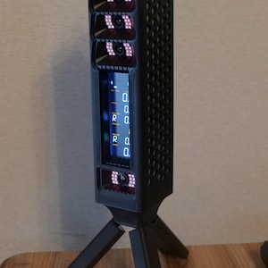 May include: A black vertical golf launch monitor with a honeycomb-patterned side, mounted on a tripod. The device features three camera lenses with red LED lights, a digital display showing numerical data, and a green light bar at the top.