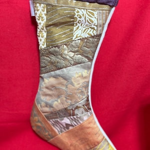 以下が含まれることがあります： パープルカラーのカフと白い裏地が付いたパッチワークのクリスマスストッキング。ストッキングは、茶色、金色、白の様々なパターンで様々な生地で作られています。