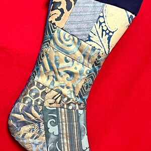 以下が含まれることがあります： 黒の折り返しと、折り返しに「手作り」と印刷された、青と金の色のパッチワークのクリスマスストッキングです。