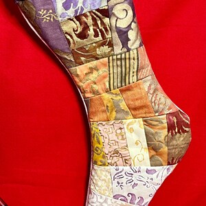 以下が含まれることがあります： パープルカフとホワイトライニングが付いたパッチワーククリスマスストッキング。ストッキングは、ブラウン、ゴールド、ピンクの色合いの花柄と幾何学模様のさまざまな生地で作られています。ストッキングには「birchhome.net」と書かれたタグが付いています。