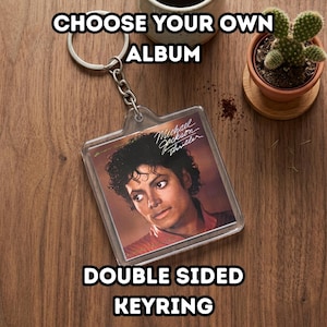 Puede incluir: Un llavero de doble cara con la portada del álbum "Thriller" de Michael Jackson. El llavero de acrílico transparente tiene un anillo y una cadena de metal plateado. El texto "CHOOSE YOUR OWN ALBUM" y "DOUBLE SIDED KEYRING" está impreso en blanco.