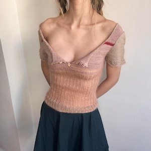 Puede incluir: Top de punto melocotón con hombros descubiertos y mangas cortas. El top presenta un escote en V con tres pequeños botones dorados. Las mangas son de color beige claro con una raya roja en la manga izquierda. La mitad inferior del top es de un color melocotón más oscuro.