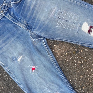 Könnte beinhalten: Eine blaue Jeans mit mehreren Patches und sichtbaren Gebrauchsspuren. Die Jeans wurde mit verschiedenen Stoffen und Nähten repariert.