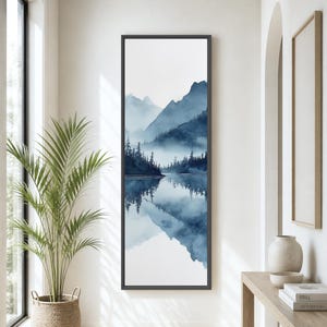 May include: A tall, vertical framed wall art piece featuring a watercolor-style landscape in shades of blue and white. The artwork depicts a misty mountain range and a dense forest reflected in a calm body of water, set within a thin black frame.