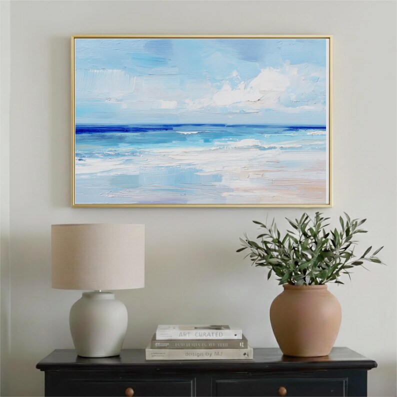a painting of a serene ocean scene hanging on a wall above a black dresser with a lamp and a vase of greenery.
