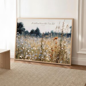 The image is a framed painting depicting a field of wildflowers, with a text overlay that reads "Be still and have that I am God".