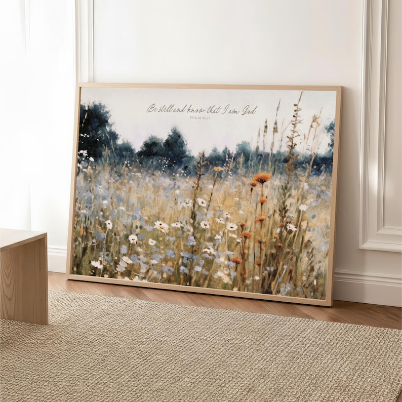 The image is a framed painting depicting a field of wildflowers, with a text overlay that reads "Be still and have that I am God".