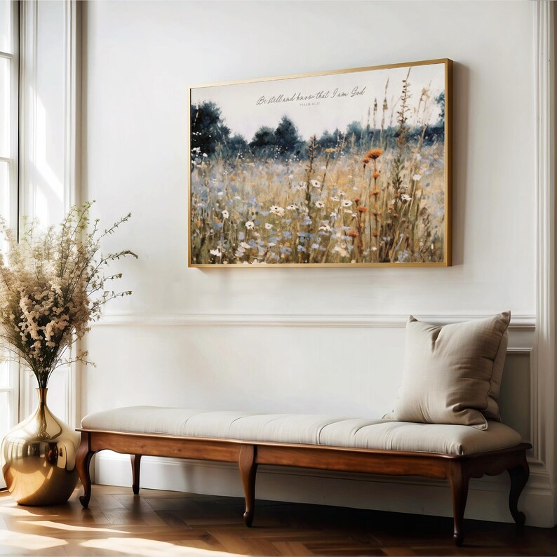 a serene indoor setting with a large painting of a field of flowers hanging on the wall above a wooden bench. The painting is framed in gold and has the text "Be still and know that I am God" written on it. The bench is positioned in front of the painting, and there is a vase of flowers on the left side of the image.