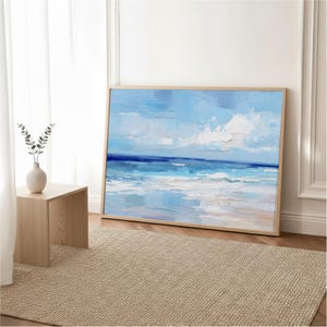 a large painting of a serene ocean landscape, with a blue sky and white clouds, hanging on a wall in a room with a wooden floor and a beige rug.