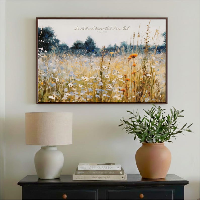 a framed painting of a field of flowers hanging on a wall above a wooden table with a lamp and a potted plant.
