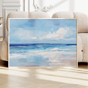 a large framed painting of a serene ocean landscape, with a blue sky and white clouds, displayed on a wooden floor in front of a beige sofa.