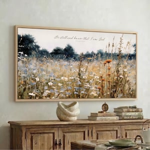 a large framed painting of a field with flowers hanging on a wall above a wooden cabinet. The cabinet has a stack of books and a small white sculpture on top of it.