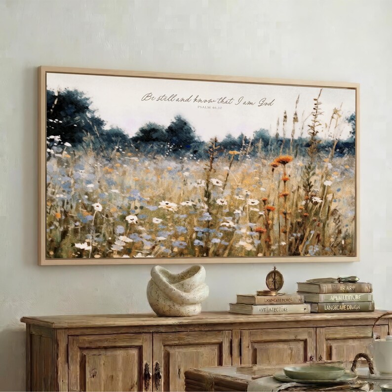 a large framed painting of a field with flowers hanging on a wall above a wooden cabinet. The cabinet has a stack of books and a small white sculpture on top of it.