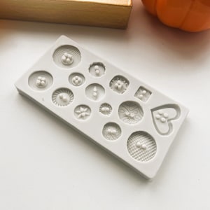 May include: A white silicone mould with 15 different shapes for making buttons. The shapes include circles, squares, hearts, and a variety of patterned designs.